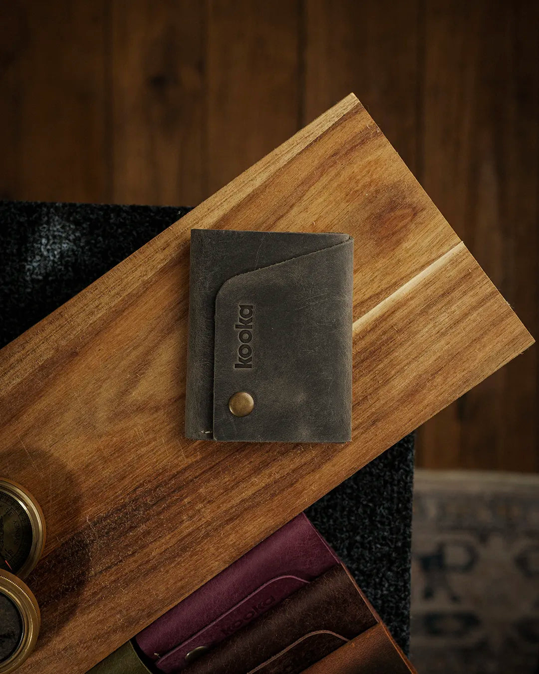 Full Grain (Üst Tabaka) Crazy Deri dikey bifold cüzdan, koyu gri-zeytin rengi doğal çekme deri yüzeyiyle; üzerindeki antik pirinç çıtçıt kapama detayı, lazer kazıma kooka markası ve angled ön kart bölmesiyle öne çıkan, el işçiliği kalitesini yansıtan kompakt bir deri cüzdan