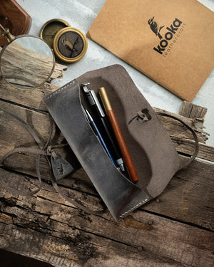 El dikişi Full Grain (Üst Tabaka) Crazy Deri kalemlik, açık pozisyonda içinde çeşitli kalemler; gri tonlu mat yüzeyde pull-up etkisi ve doğal doku izleri belirgin. Kooka Leathercraft Yatay Kalemlik, sol tarafta suet çekim kulakçığı ve gevşek bırakılmış deri bağcıklı kapama sistemiyle; kenar boyunca düzgün krem renkli el dikişi ve kapak üzerinde kabartma kookabura logosu detayıyla öne çıkan el yapımı deri kırtasiye aksesuarı.