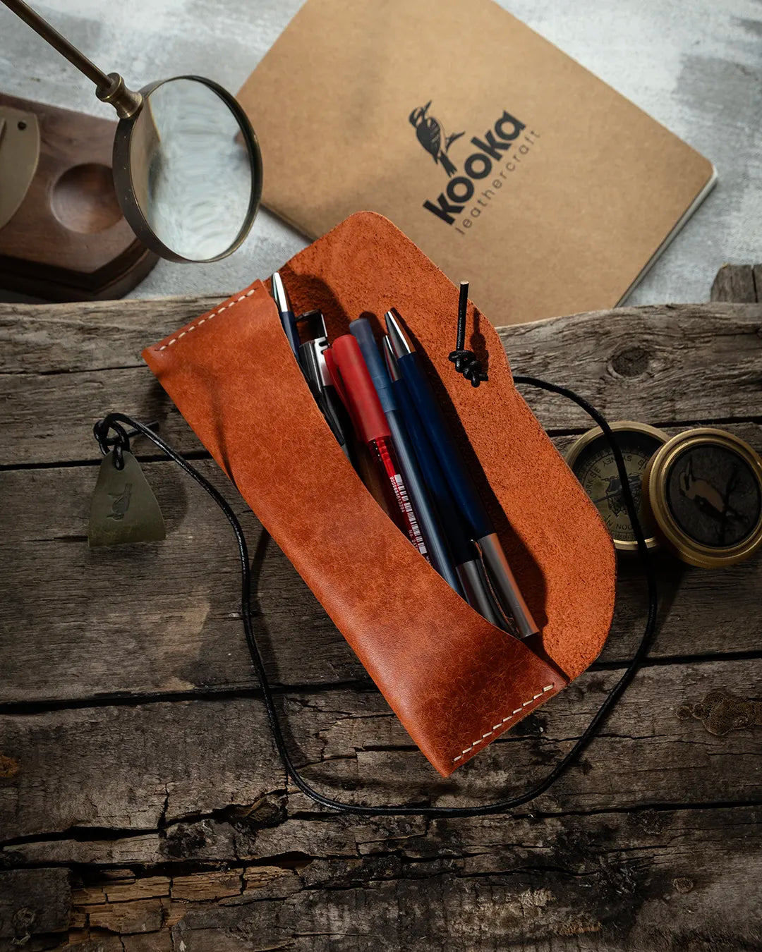 El dikişi deri kalemlik, taba rengi Pueblo derisiyle hazırlanmış KOOKA Leathercraft imzalı yatay kalemlik açık konumda, içinde dolma kalem ve tükenmez kalemlerle; suet iç yüzey, siyah deri kordon ve kookaburra logolu üçgen çekme askısıyla tam görünüm.