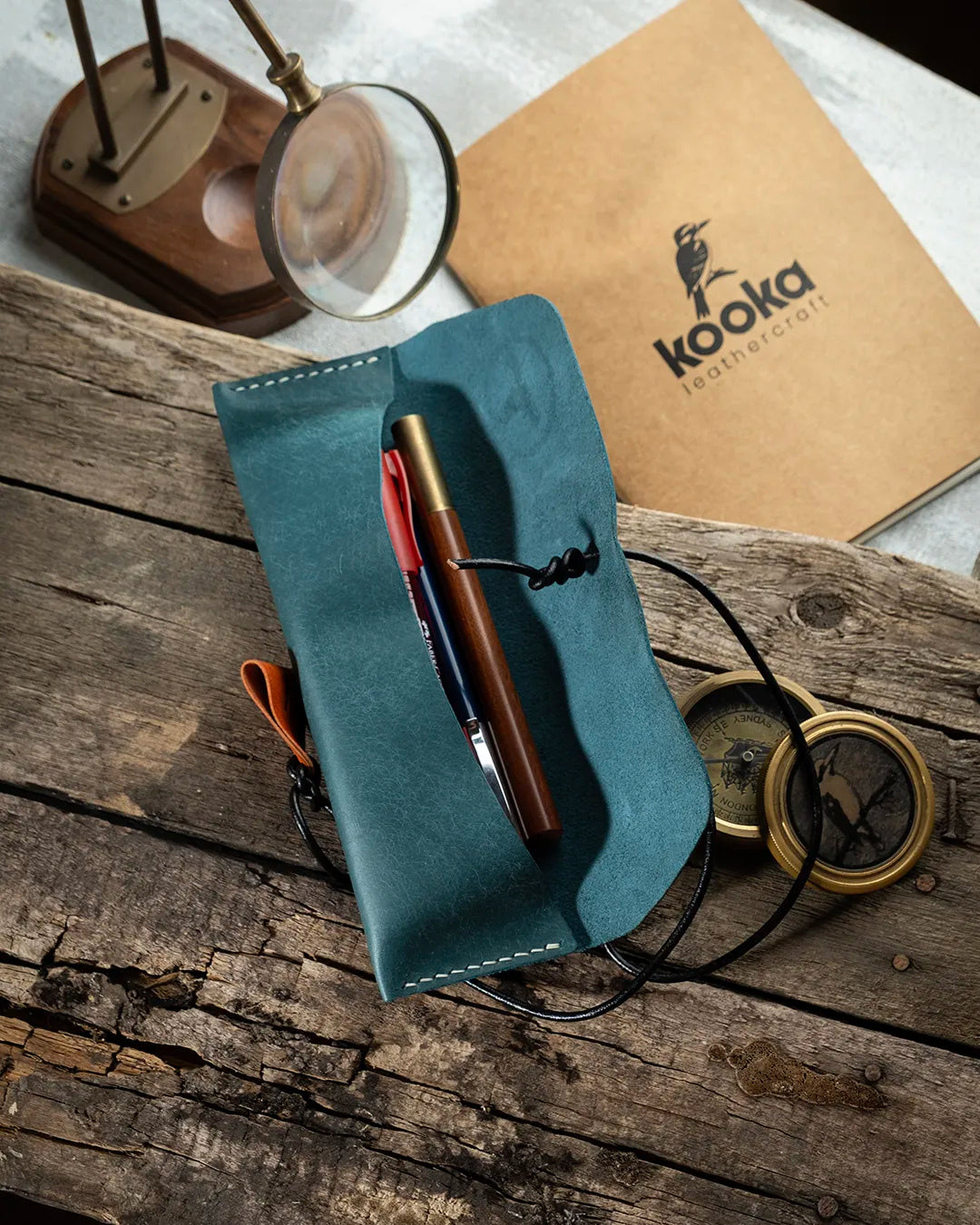 El dikişi deri kalemlik, mavi Pueblo deri yüzeyiyle zengin bir doku ve doğal çekme efekti sunar; açık konumda içindeki kalemlerle birlikte sergilenen KOOKA Leathercraft imzalı bu yatay model, turuncu süet çekme kulpu ve dokuma bağlama ipi detaylarıyla özgün el işçiliğini yansıtır.