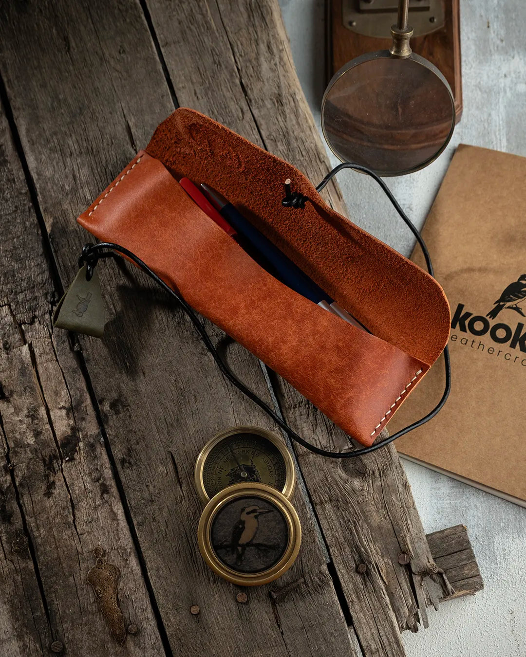 El dikişi deri kalemlik, taba rengi Pueblo derisiyle hazırlanmış KOOKA Leathercraft imzalı bu yatay kalemlikte açık konumuyla iç bölme ve suet iç yüzey net biçimde görülmektedir; kırmızı ve lacivert kalemler barındıran poş, dışa kıvrılmış kapağıyla el işçiliği kalitesini ve sarmal kordon bağcık sistemini sergiliyor.
