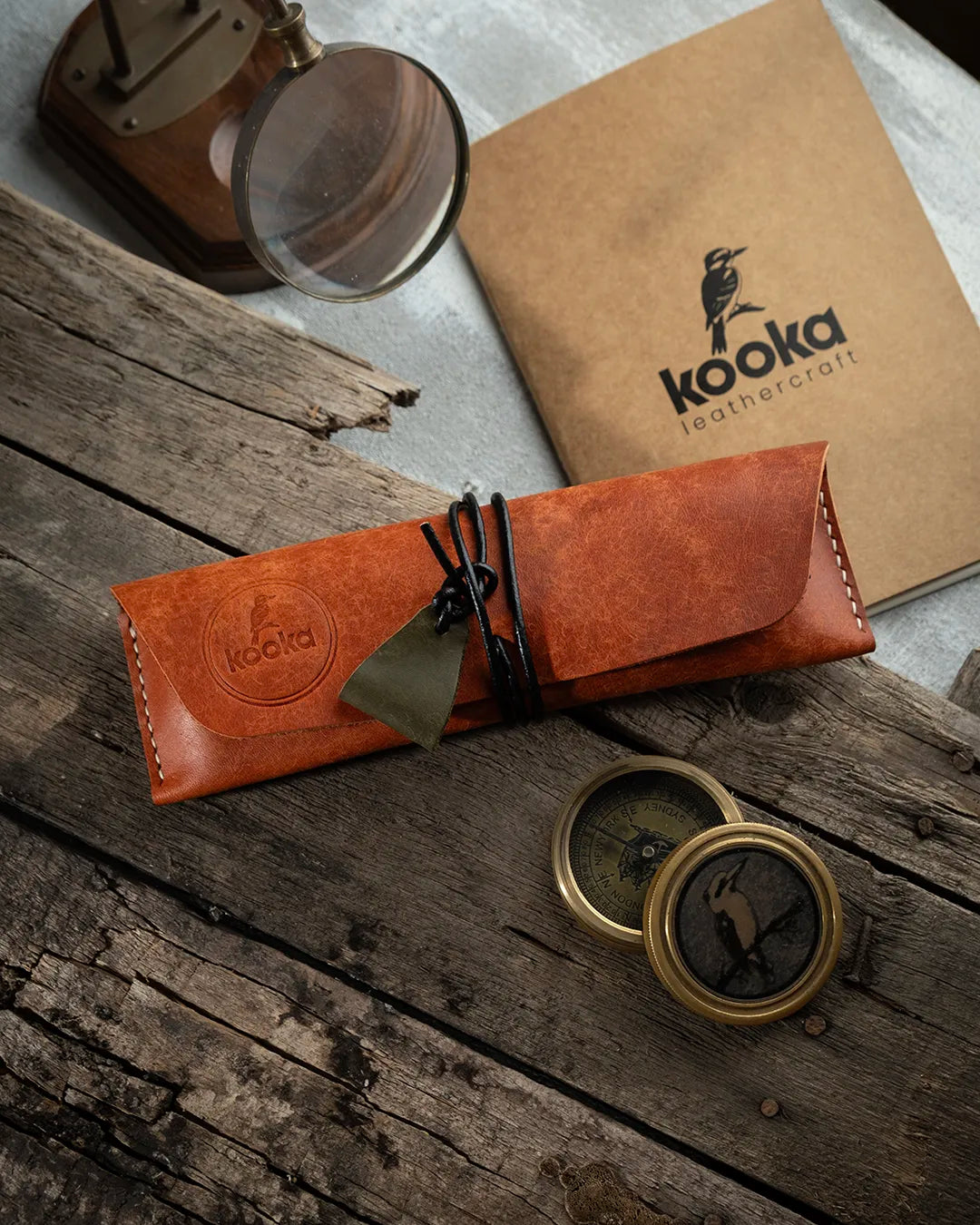 El dikişi deri kalemlik, taba renkli PUEBLO derisiyle üretilmiş, sarılı ip kilidli ve küçük üçgen süet çekme askılı kapalı konumda; Full Grain (Üst Tabaka) yüzeyin sıcak cognac tonu ve dairesel KOOKA marka damgası öne çıkıyor.