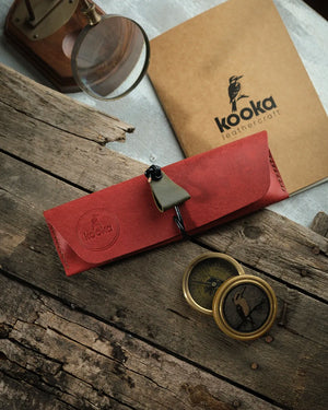 El dikişi deri kalemlik, kırmızı Pueblo derisiyle üretilmiş KOOKA Leathercraft Yatay Kalemlik, kapalı ve kordon bağlı konumda; zeytin yeşili üçgen süet çekme kulpu ile kontrast oluşturan daire içi kookaburra kuşu kabartma marka damgası ve bej el dikişi detayları öne çıkıyor.