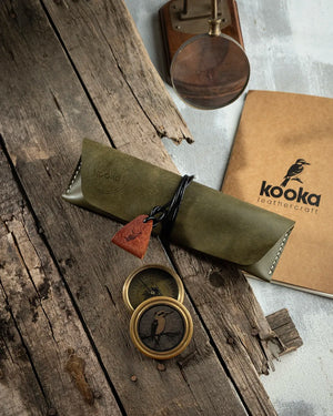 El dikişi PUEBLO deri kalemlik, zeytin yeşili rengi ve mat kadifemsi yüzey dokusuyla öne çıkan Kooka Leathercraft imzalı tam kapamalı tasarım; sargılı deri kordon kilidi ve bronz rengi üçgen çekme askısıyla tamamlanan, Full Grain (Üst Tabaka) derinin doğal karakterini yansıtan el yapımı kalem çantası.