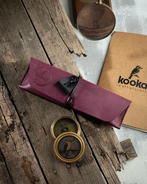El dikişi mor Pueblo deri kalemlik, KOOKA Leathercraft imzasıyla; kapaklı sarma tasarımı, siyah deri üçgen çekme askısı ve dairesel kabartma marka damgasıyla öne çıkan butik el işçiliği kalem çantası
